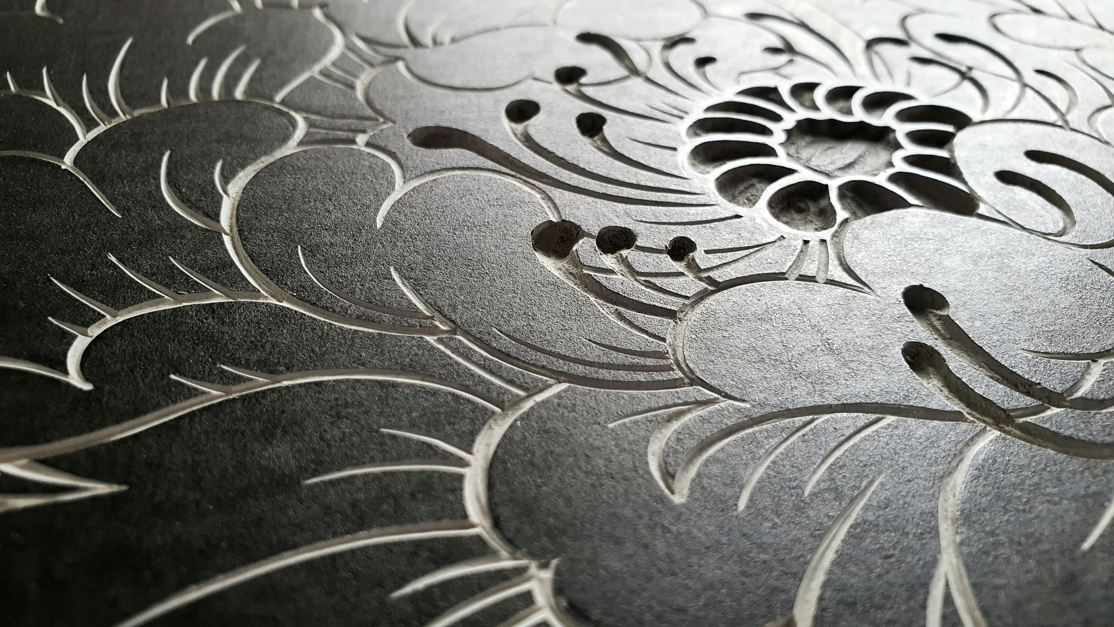 A hand-carved linocut block. Close-up of a tattoo-style black peony flower shown from an angle. Light reflecting from the block.