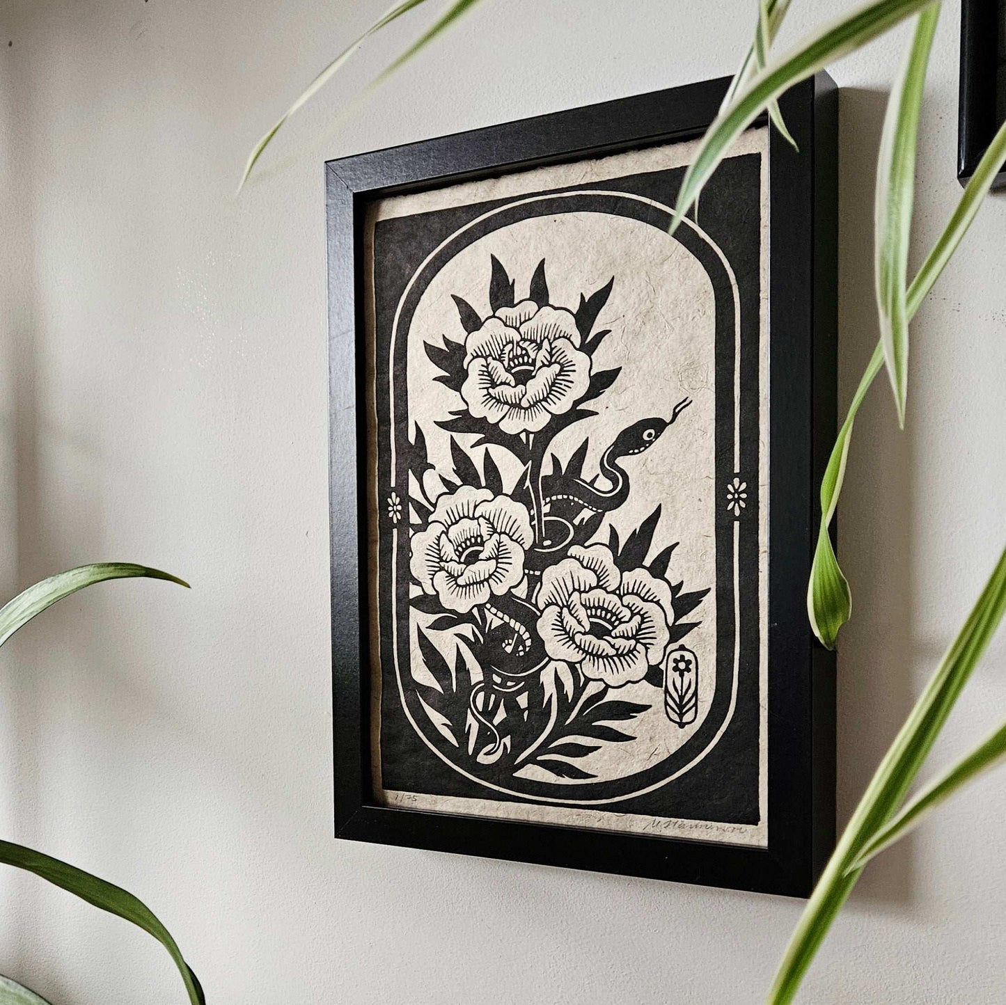 A framed woodcut print of a snake and flowers hanging on on a wall