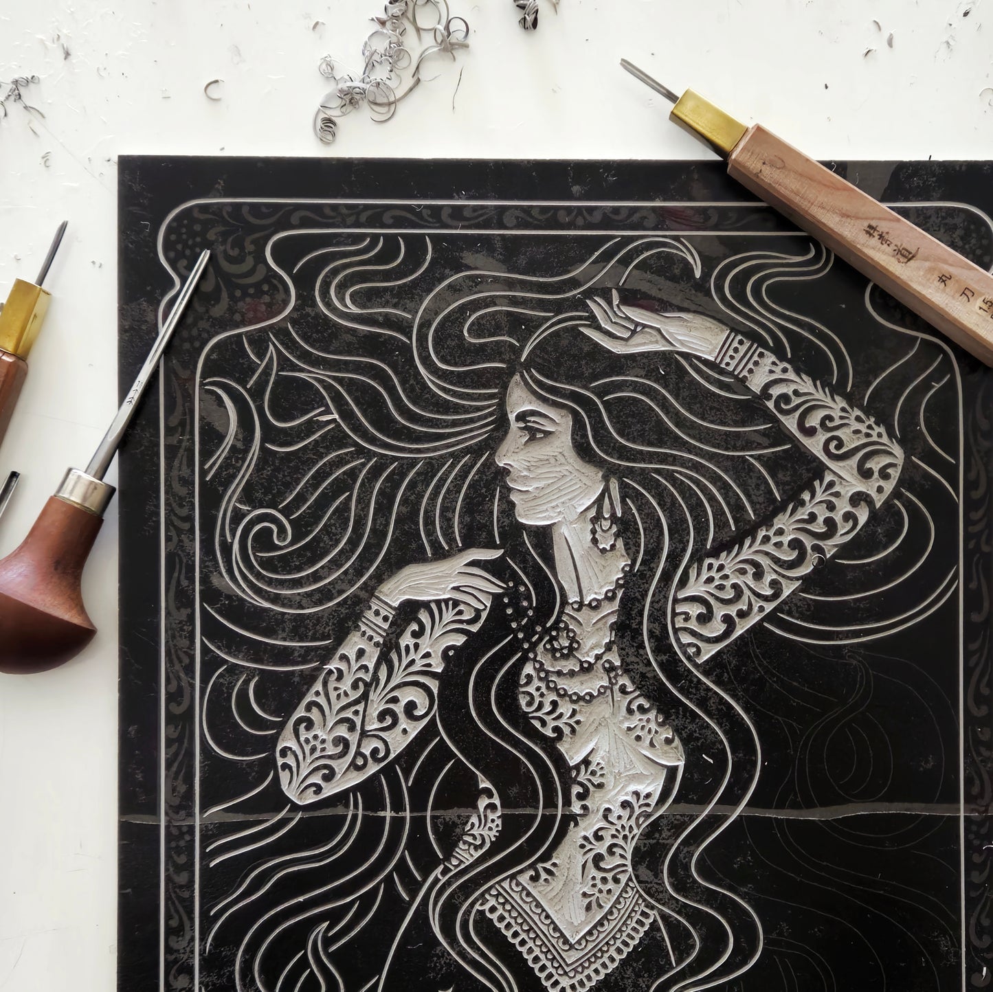 Artistic engraving of a woman with detailed patterns on black surface, surrounded by engraving tools.