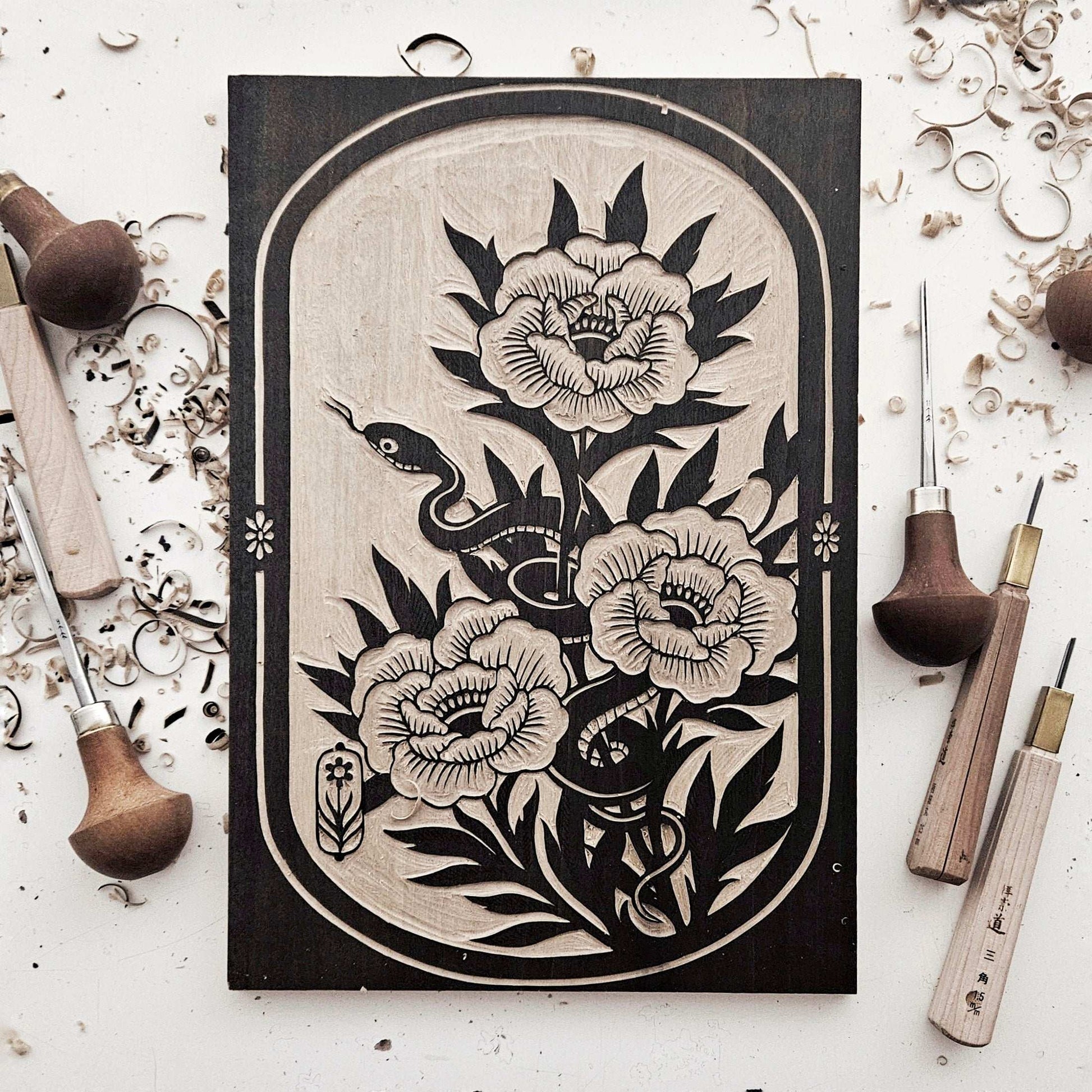 Hand carved woodcut printing block of a snake and flowers surrounded by woodcarving tools and wood shavings