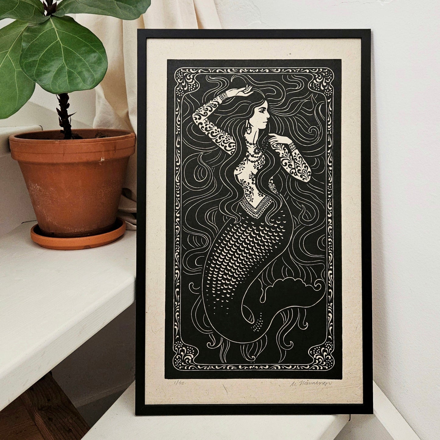 Framed artwork of a mermaid with intricate patterns on a white surface next to a potted plant.