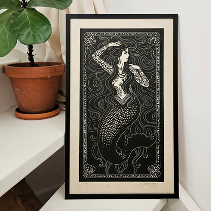 Framed artwork of a mermaid with intricate patterns on a white surface next to a potted plant.